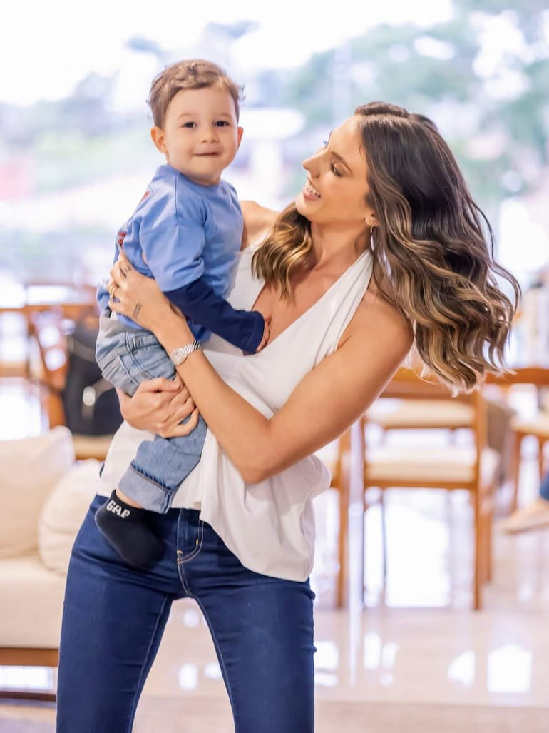 Mujer vestida con blusa blanca y jeans ajustados sonríe mientras sostiene a un niño pequeño con camiseta azul en un entorno hogareño.