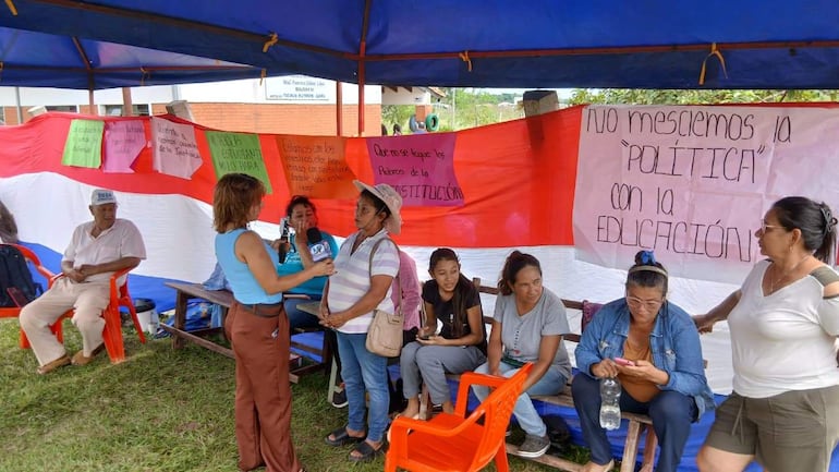 Mujer con micrófono entrevista a otra mujer en camiseta clara y sombrero, mientras se observan carteles sobre educación.