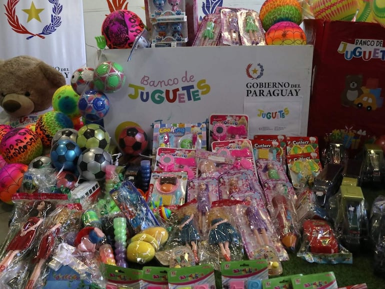 Variedad de juguetes incluyendo pelotas, muñecas y juegos de mesa en un entorno festivo, con cartel del Banco de Juguetes.