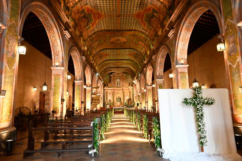 Interior de la iglesia con luz suave, altos arcos decorados y altar adornado con flores blancas, sin personas visibles.