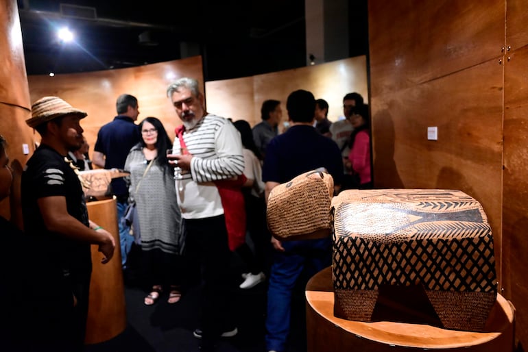 Hombre con sombrero y camiseta negra conversa, mientras mujer de cabello largo lleva bolsa en ambiente con exhibiciones artísticas.