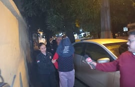 Mujer con uniforme oscuro sostiene a hombre en capucha, mientras otro hombre con micrófono observa, en un entorno urbano nocturno.