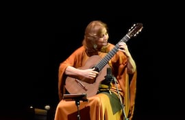 Berta Rojas sentada, tocando guitarra con túnica naranja, en un escenario elegante y oscuro.