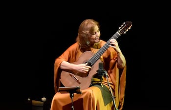 Berta Rojas sentada, tocando guitarra con túnica naranja, en un escenario elegante y oscuro.