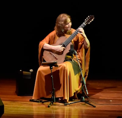Berta Rojas sentada, tocando guitarra con túnica naranja, en un escenario elegante y oscuro.