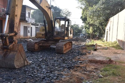 Excavadora amarilla en acción sobre terreno pedregoso, rodeada de edificios y vegetación en un entorno urbano.