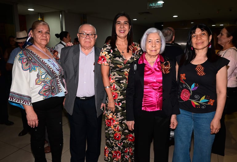 Mujeres con vestimentas decoradas y un hombre canoso con gafas, sonriendo en un entorno social con iluminación tenue.