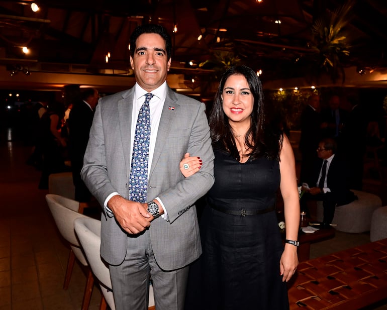 Hombre sonriente en traje gris junto a mujer con vestido negro en un evento formal, rodeados de un ambiente festivo.