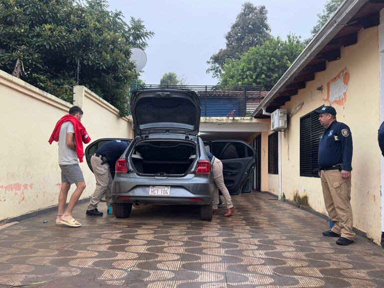 Tres hombres presentes: uno con toalla roja, otro agachado junto al auto gris y un agente con uniforme en garaje residencial.