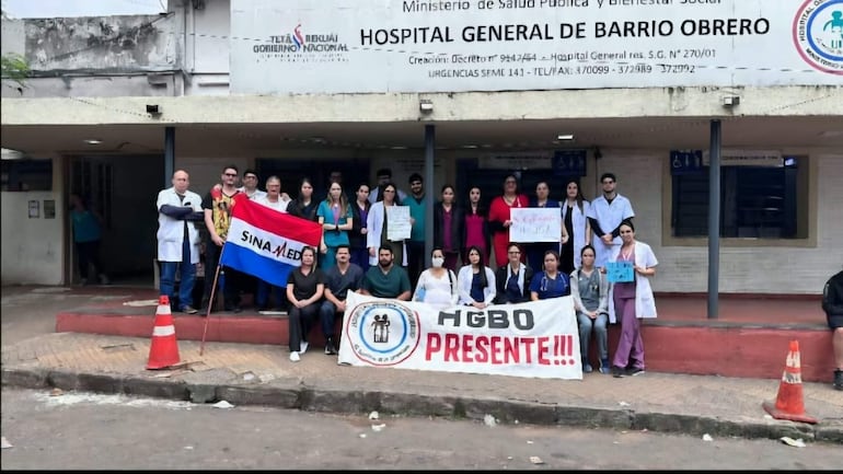Grupo de médicos en uniformes médicos sostiene carteles y bandera paraguaya frente al Hospital General de Barrio Obrero.