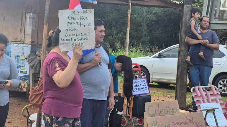 Mujer con cartel en mano y hombre a su lado, mientras otros miran teléfonos. Vehículos y bandera paraguaya en el fondo.