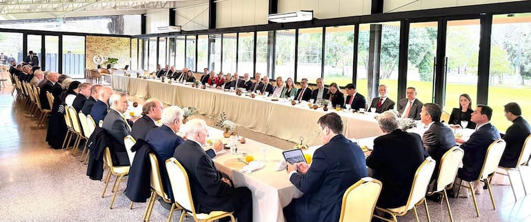 Grupo de personas en mesa elegante, conversando con ambiente iluminado por luz natural, decoraciones simples y smartphones visibles.