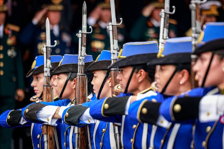Seis mujeres uniformadas en azul sostienen rifles, en un acto militar con hombres saludando al fondo.