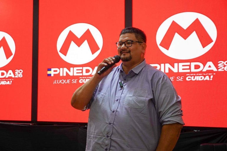 Hombre robusto con gafas sostiene un micrófono, fondo rojo con 'PINEDA 2026' y '¡LUQUE SE CUIDA!' en pantallas.
