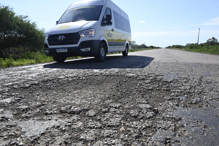 Camioneta blanca con detalles en amarillo detenida en una carretera llena de baches, rodeada de vegetación baja.