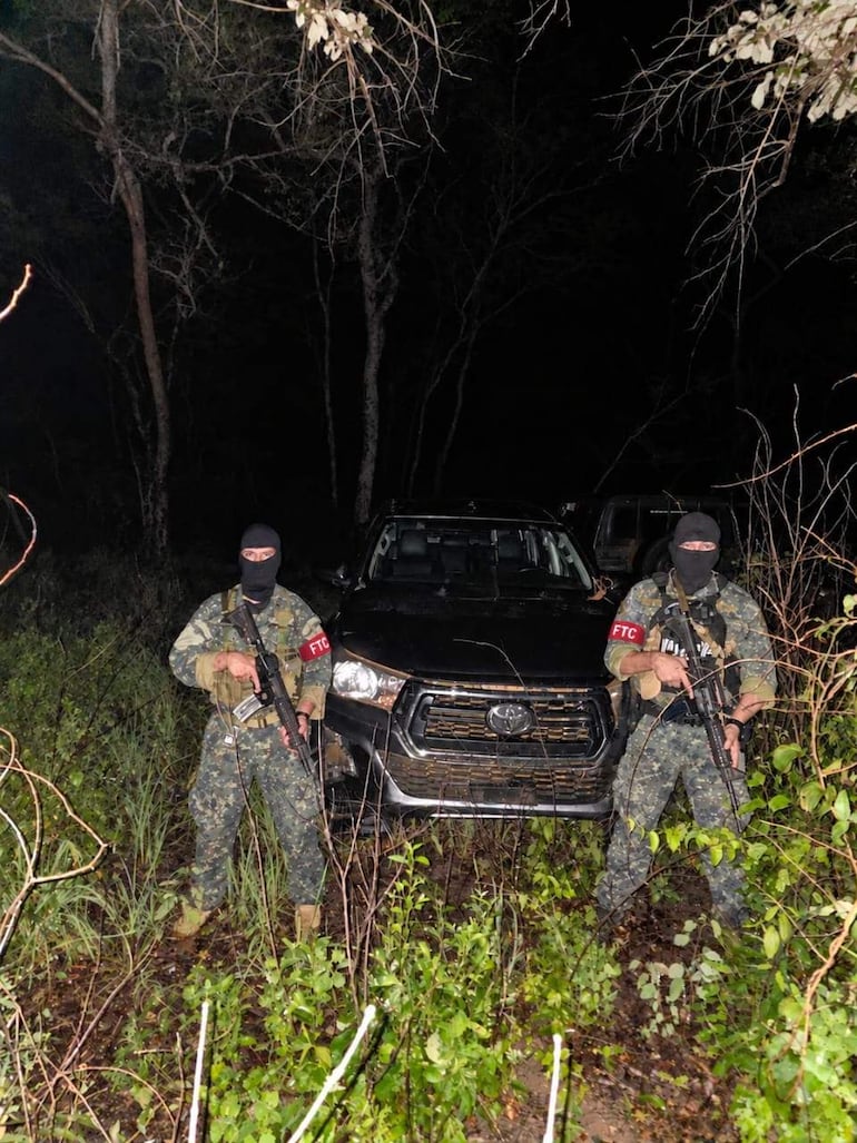 Dos hombres armados con uniformes de camuflaje y pasamontañas, junto a un SUV Toyota, en un entorno natural oscuro.