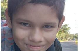Niño de cabello corto y oscuro, sonriente, viste camiseta oscura y mochila colorida, rodeado de árboles en un camino.