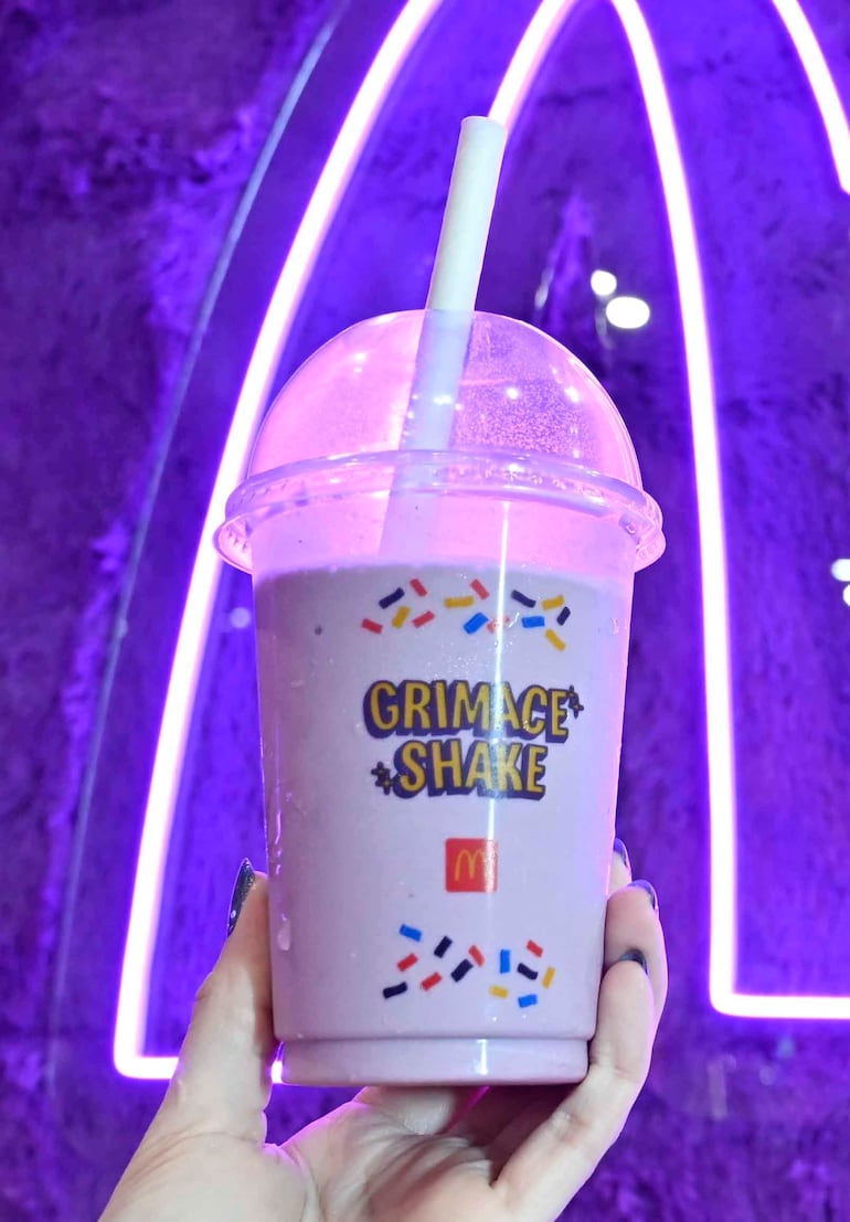 Vaso de Grimace Shake sostenido con una mano, fondo iluminado en tonos púrpuras y logo de McDonald's visible.