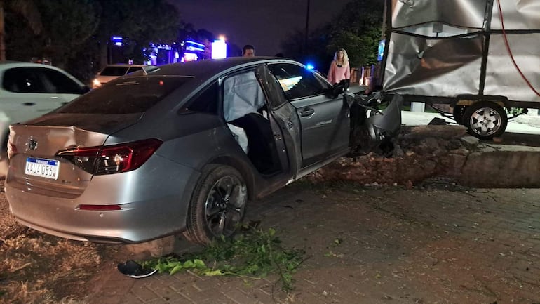 Automóvil gris dañado en la acera con personas observando el incidente, bajo iluminación artificial.