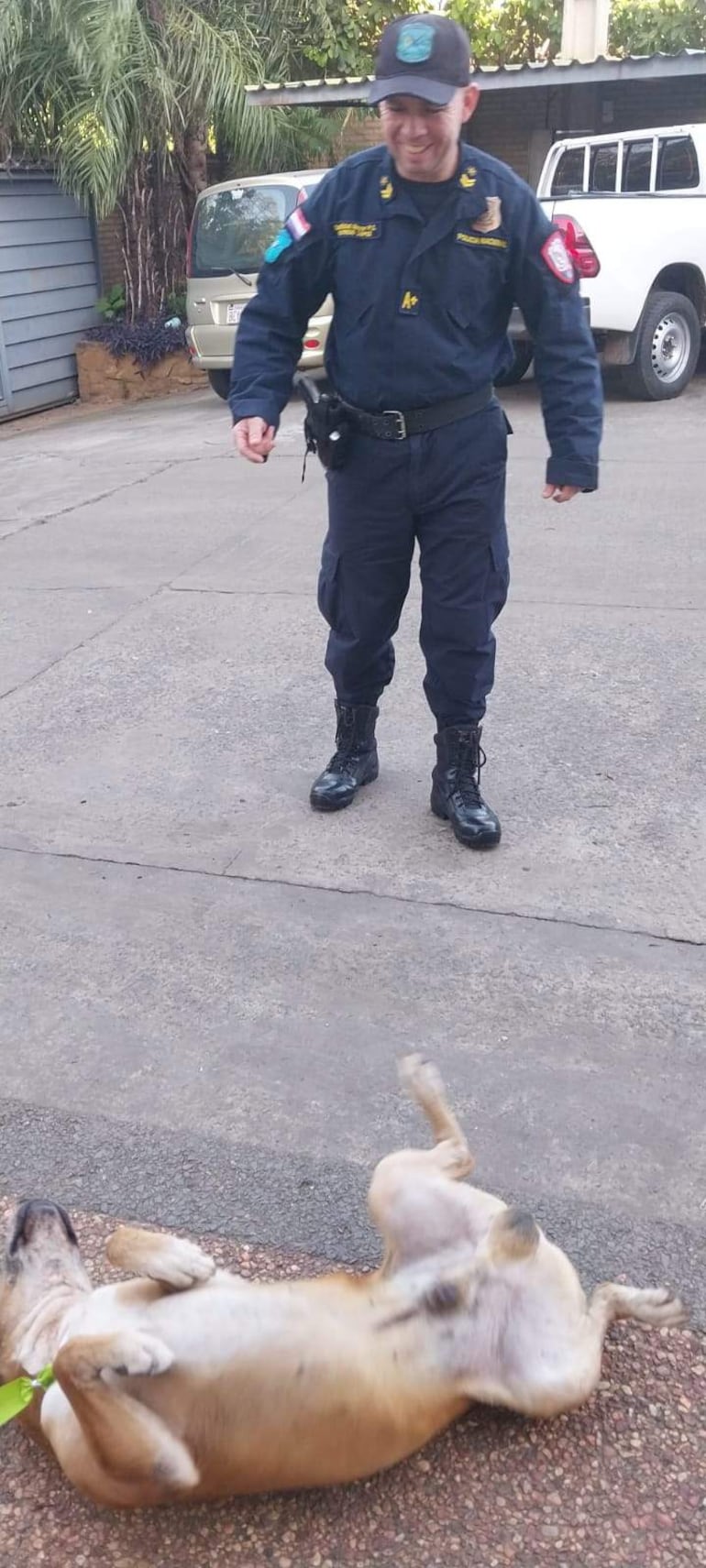 Oficial de policía con uniforme azul oscuro sonríe mientras un perro marrón juega en el suelo.