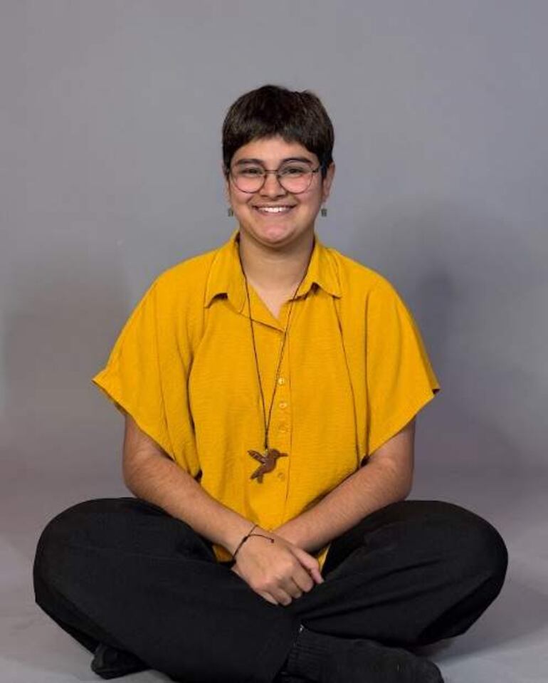 Hombre sonriente con gafas, vestido con camisa amarilla y pantalones oscuros, sentado en el suelo en pose relajada.