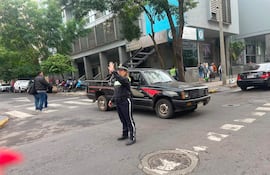 Oficial en uniforme levantando la mano en señal de alto, con un hombre a su izquierda y vehículos en la calle de fondo.