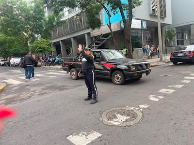 Oficial en uniforme levantando la mano en señal de alto, con un hombre a su izquierda y vehículos en la calle de fondo.