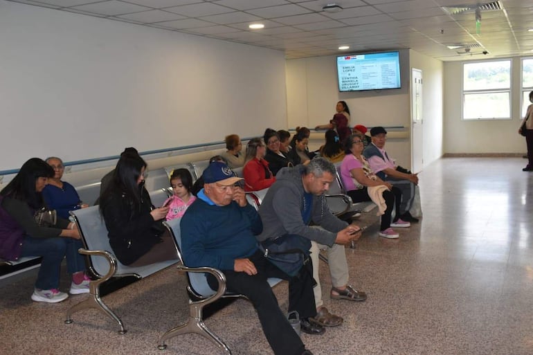 Varias personas en el hospital, algunos usan móviles, un hombre con gorra azul se sienta en el centro junto a un adulto mayor.