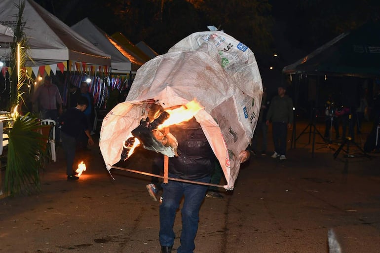Hombre con abrigo oscuro y jeans carga un muñeco en llamas en un ambiente festivo iluminado por luces y decoraciones.