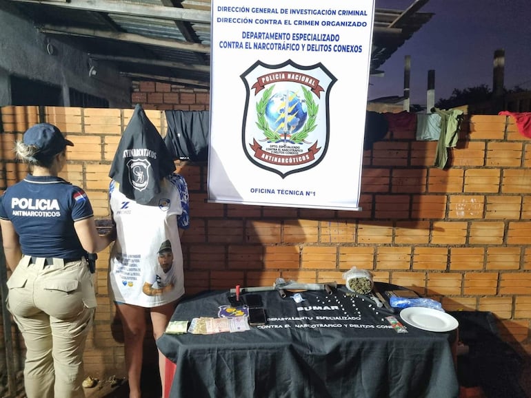 Mujer con camiseta de 'Antinarcóticos' asiste a detenido con capucha negra, en operativo policial rodeado de objetos y dinero.