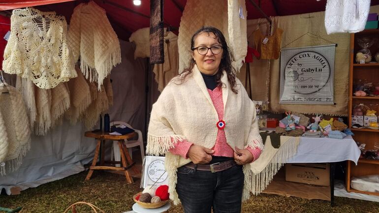 Lucía Jaquet sonríe mientras presenta su chal de lana, con fondo de productos artesanales y un cartel que anuncia su negocio.