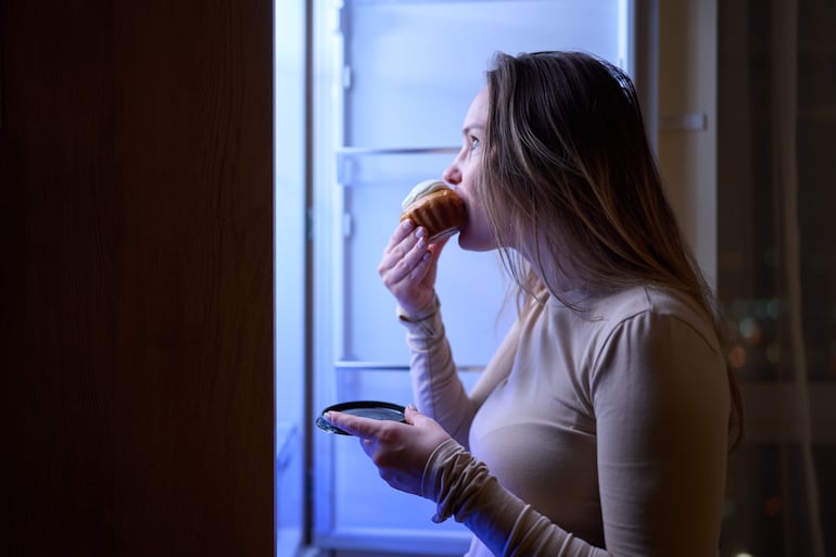 Mujer come frente a la heladera de noche.