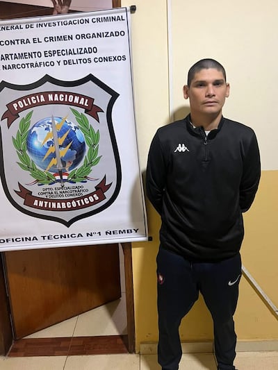 Hombre de pie en sudadera oscura, con logo de Kappa, en oficina con cartel de Policía Nacional, expresión neutral.