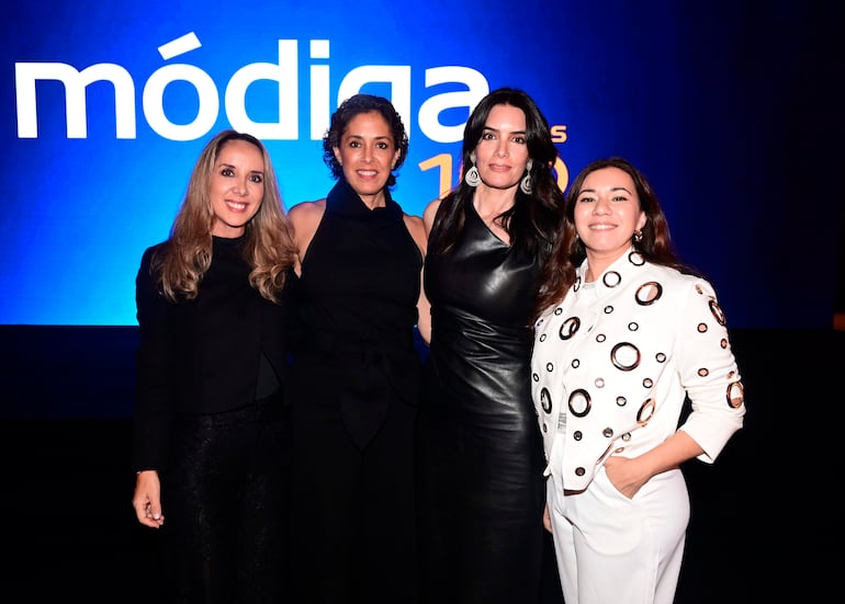 Cuatro mujeres se agrupan sonriendo: una con blusa negra, otra en vestido negro, una más en vestido de cuero; fondo azul con 'módiga'.