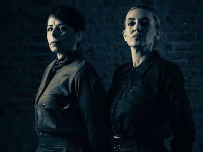 Dos mujeres en ropa oscura, una con cabello corto y expresión seria, y la otra con cabello recogido y mirada intensa, posan en un ambiente oscuro.