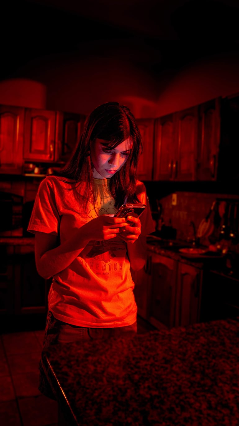 Joven de cabello largo y oscuro, con camiseta clara, revisa su teléfono en cocina oscura con luz roja.