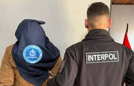 Dos hombres en interior, uno cubierto con pañuelo de INTERPOL y otro con chaquetón de la misma organización, ambiente serio.