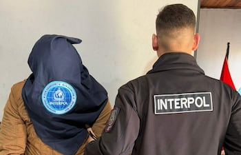Dos hombres en interior, uno cubierto con pañuelo de INTERPOL y otro con chaquetón de la misma organización, ambiente serio.