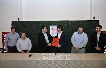 Hombres en traje oscuro se dan la mano, con un documento rojo entre ellos, en un evento formal decorado con paredes verdes.