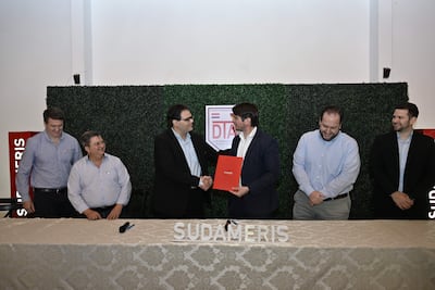Hombres en traje oscuro se dan la mano, con un documento rojo entre ellos, en un evento formal decorado con paredes verdes.