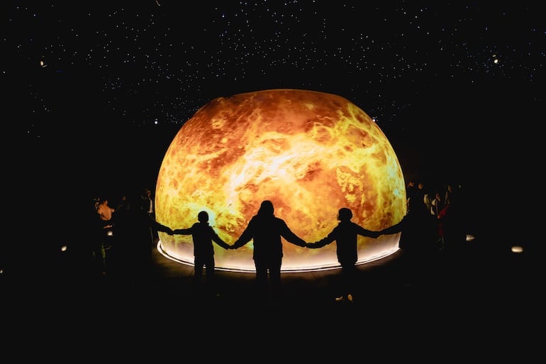 Seis hombres en un ambiente oscuro sosteniendo manos alrededor de un planeta iluminado en un evento de ciencias.