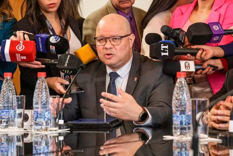 Óscar Lovera, calvo y con gafas, en traje oscuro y corbata azul, habla con varios micrófonos frente a él.
