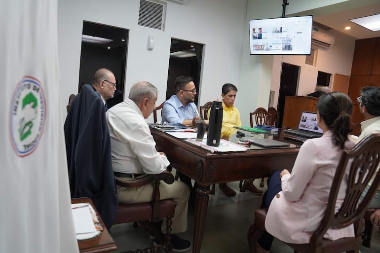 Hassel Jimmy Jiménez en camisa celeste, habla mientras otros escuchan atentos en sala de conferencias, con documentos y pantalla visible.