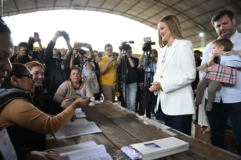 Soledad Núñez con abrigo blanco viste sonriente mientras conversa, acompañada por Bruno Defelippe y otras personas en un ambiente de votación.