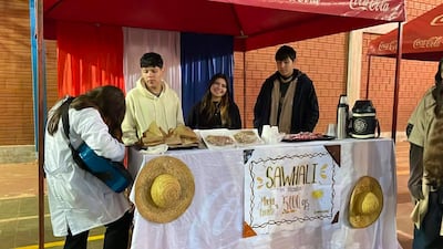 Mujer sonriente en el centro con abrigos observa a un hombre de espaldas tocando guitarra en un ambiente festivo con colores de la bandera paraguaya.