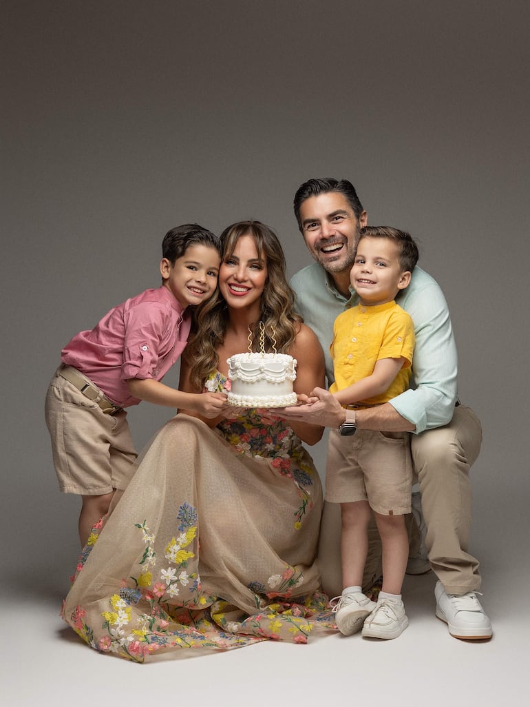 Una mujer sonriente en vestido floral sostiene un pastel, acompañada de dos niños y un hombre con barba.