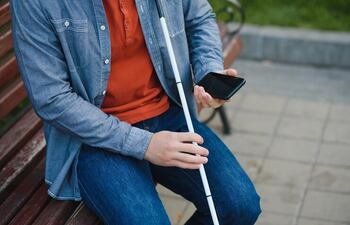 Una persona sentada en un banco público, que tiene un bastón blanco en una de sus manos y en la otra sostiene un teléfono celular.