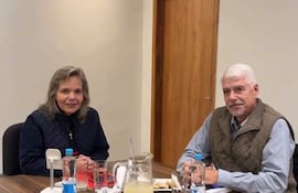 Lilian Samaniego con suéter negro y chaleco sonriente, junto a Arnoldo Wiens con chaleco gris, conversan en una mesa con bocadillos.