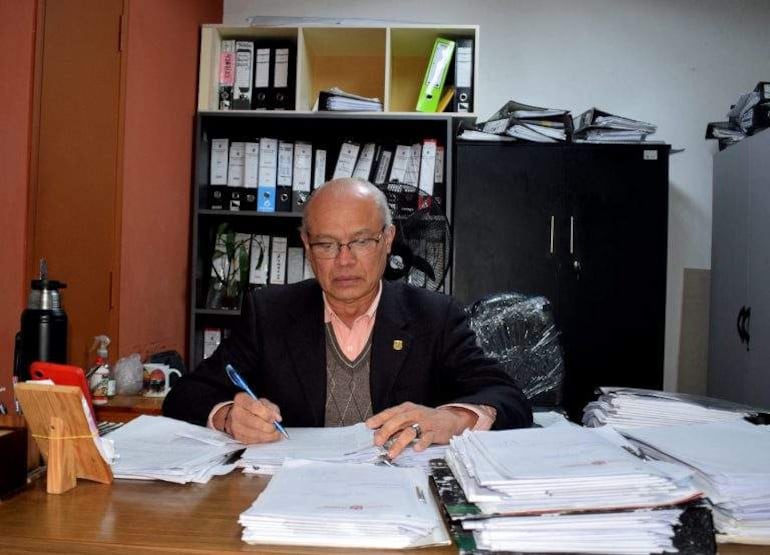 Hombre canoso con lentes y suéter gris escribe en un escritorio desbordado de documentos en su oficina.