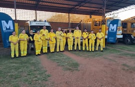 Grupo de bomberos en trajes amarillos, uno sostiene un reconocimiento, con vehículos de bomberos y pantalla publicitaria detrás.